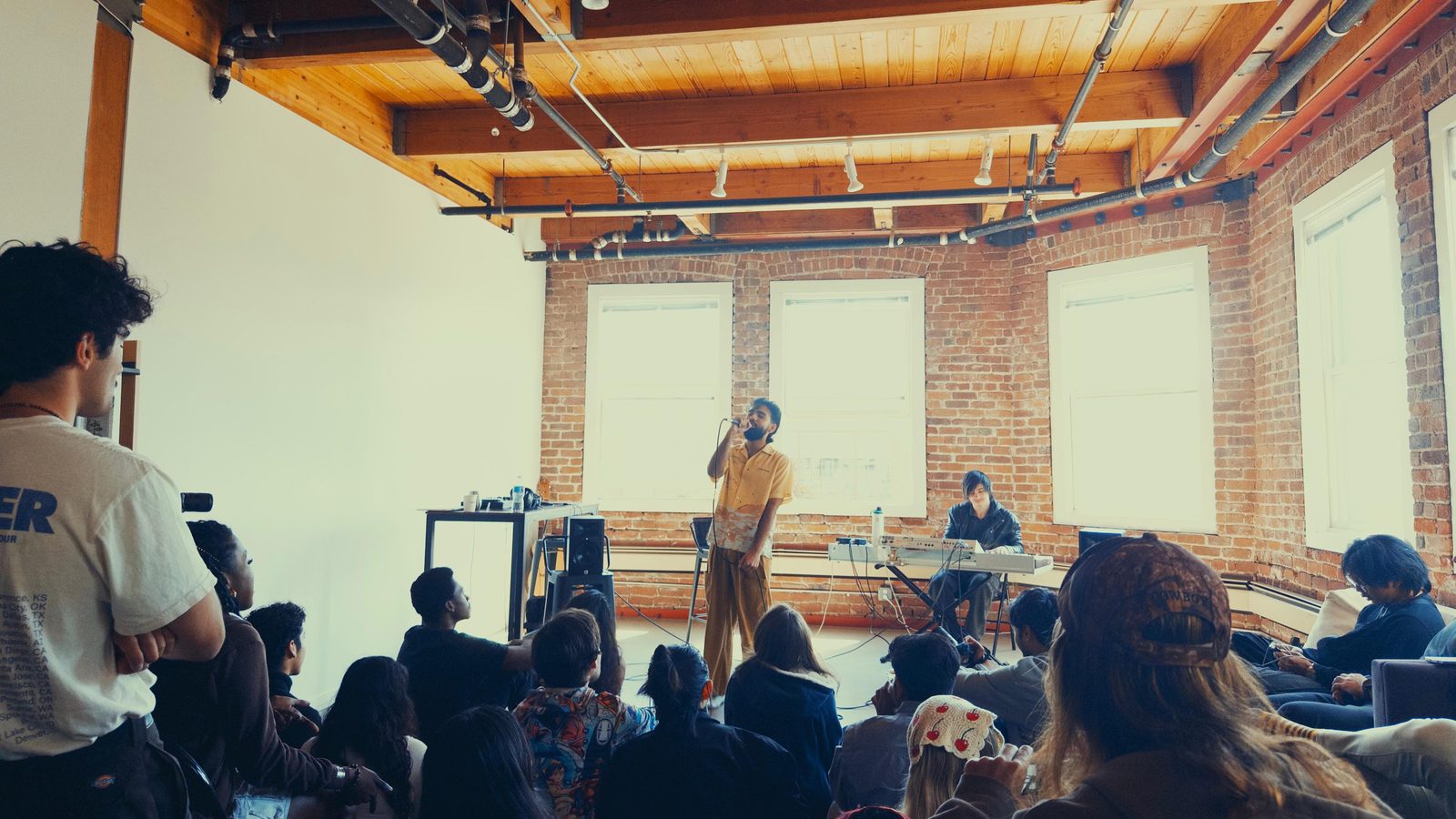 Sommaiya Angrish singing into a microphone at an intimate show in a brick-walled San Francisco loft in 2025, with a keyboardist beside him and the audience seated on the floor