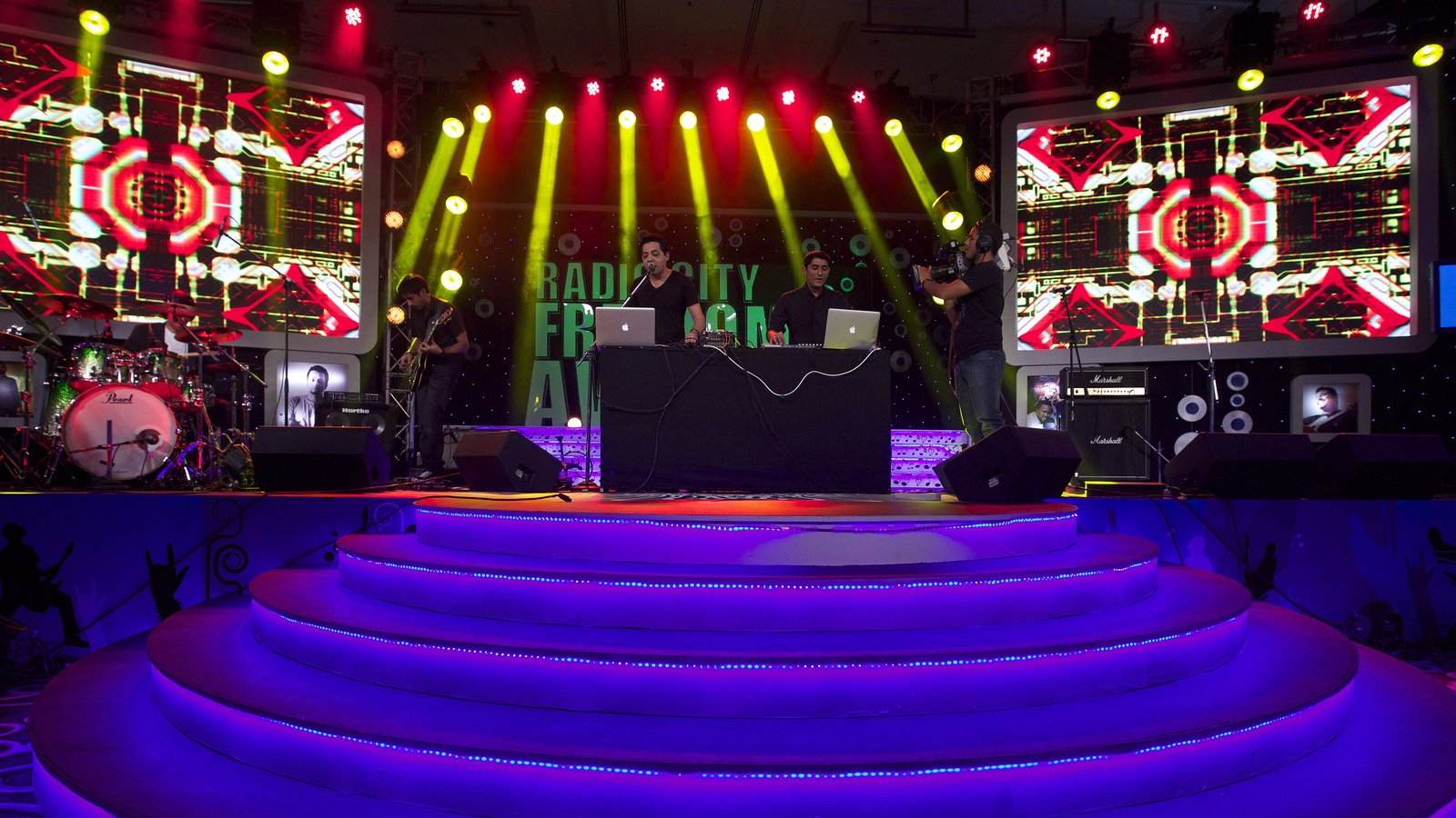 MIDIval Punditz performing behind laptops on a stage lit with yellow beams and red LED screens at the Radio City Freedom Awards in 2013