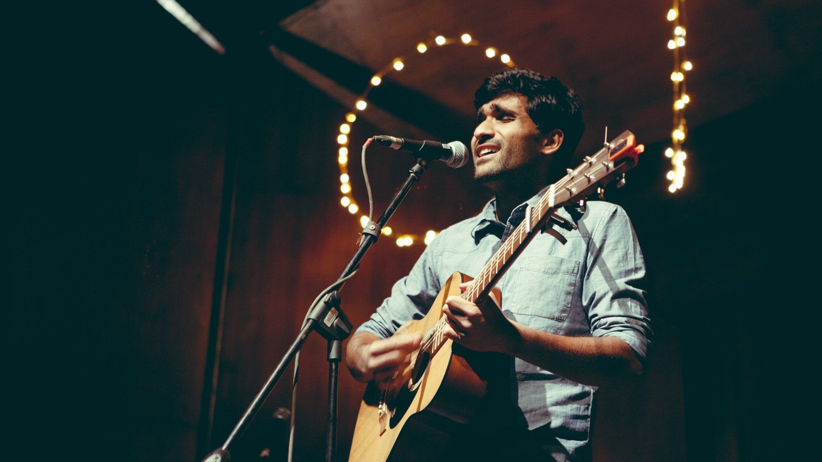 Prateek Kuhad singing into a microphone and playing acoustic guitar on a dim stage lit by string lights