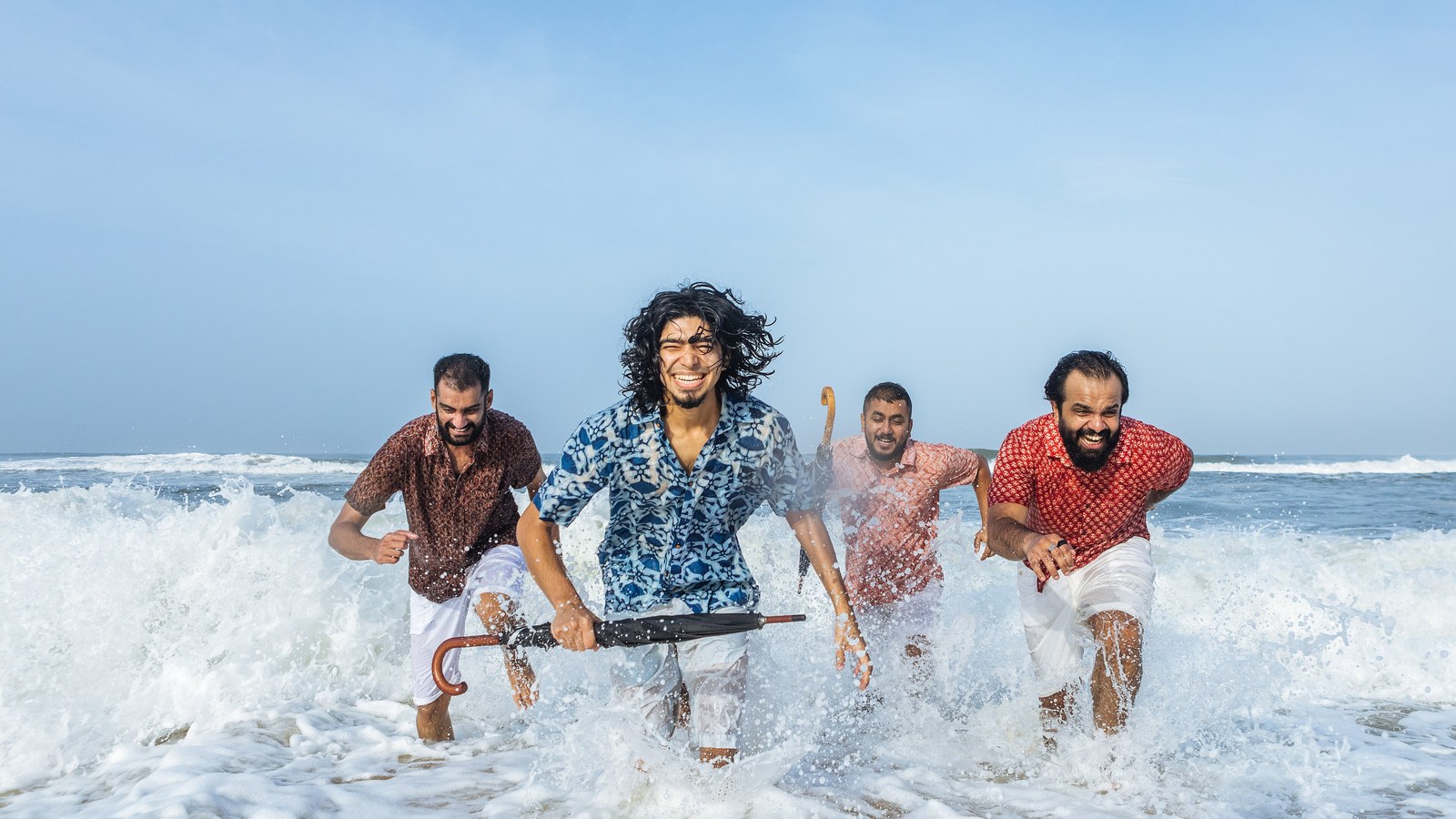 When Chai Met Toast band members running out of the sea waves on a beach, laughing, one holding an umbrella