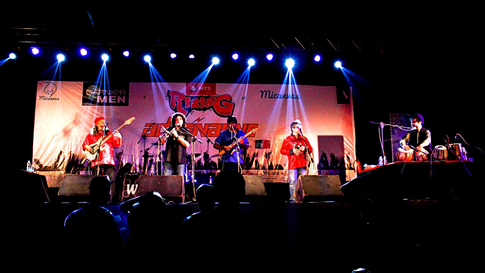 Indian Ocean perform live on a stage lit by blue spotlights, with guitarists, a flautist and a tabla player in front of a festival banner