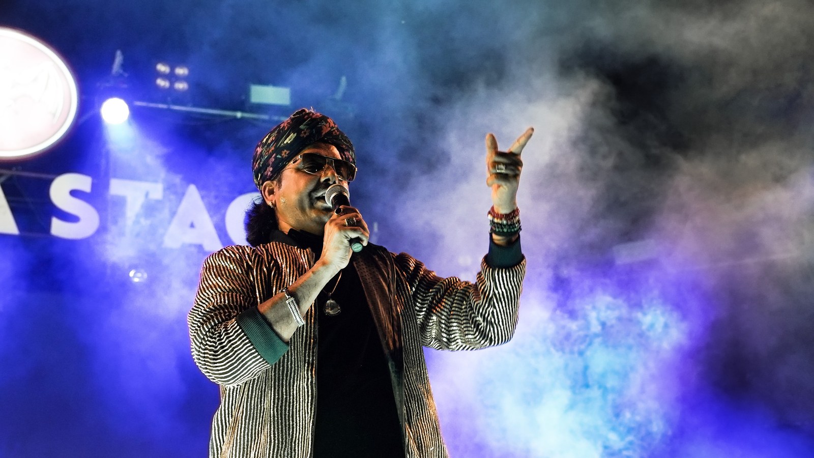 Rajasthani folk singer Mame Khan in a turban and sunglasses sings into a microphone on a smoky blue-lit stage