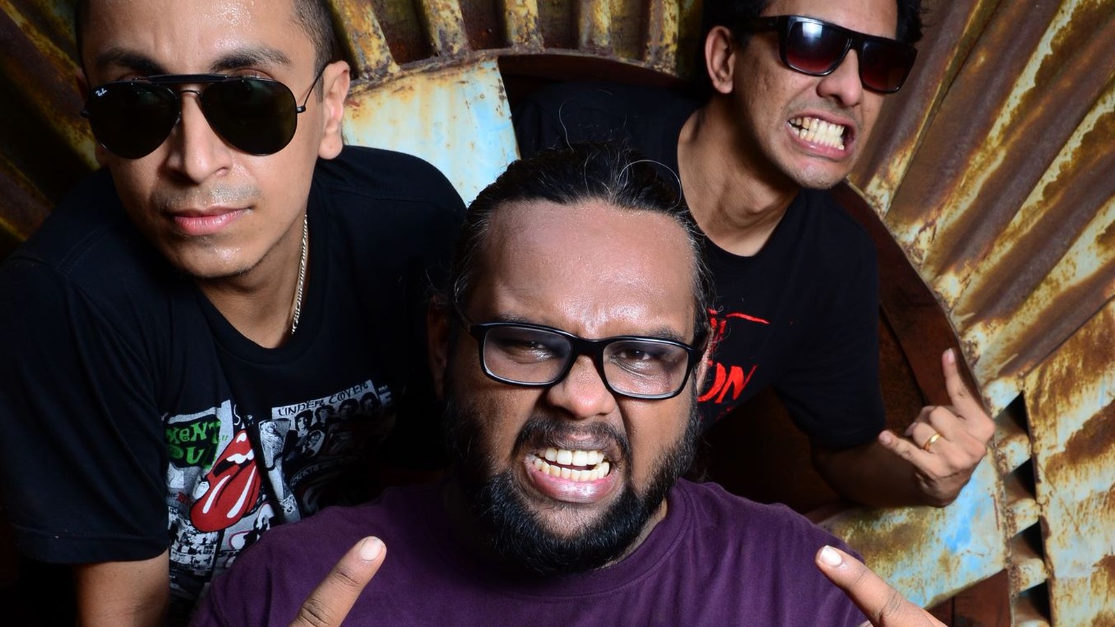 The three members of Mumbai punk band Tripwire pulling faces and making the sign of the horns in front of rusted corrugated metal