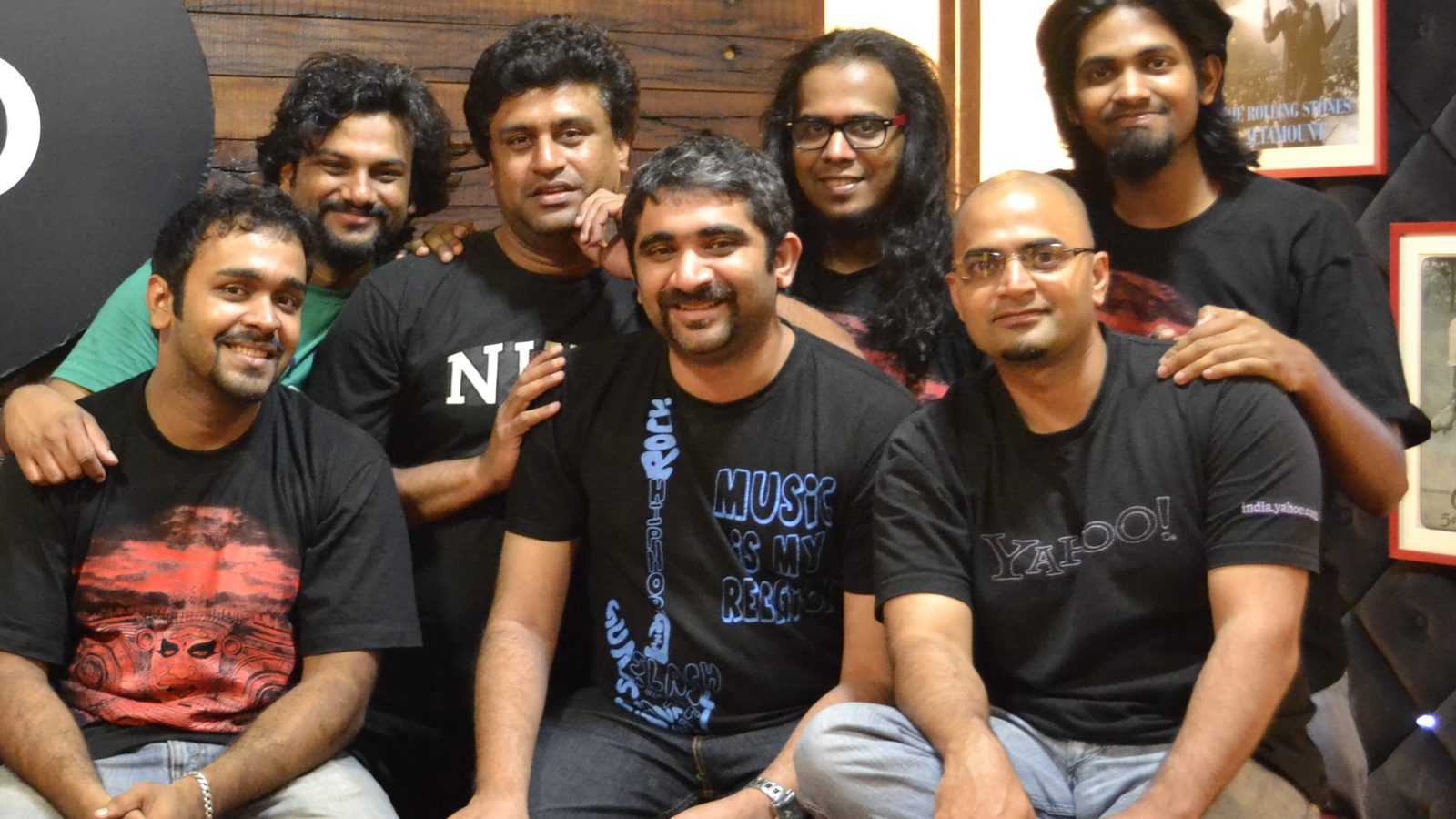 Seven members of Bengaluru band Agam smiling together in a studio with wooden walls and framed pictures