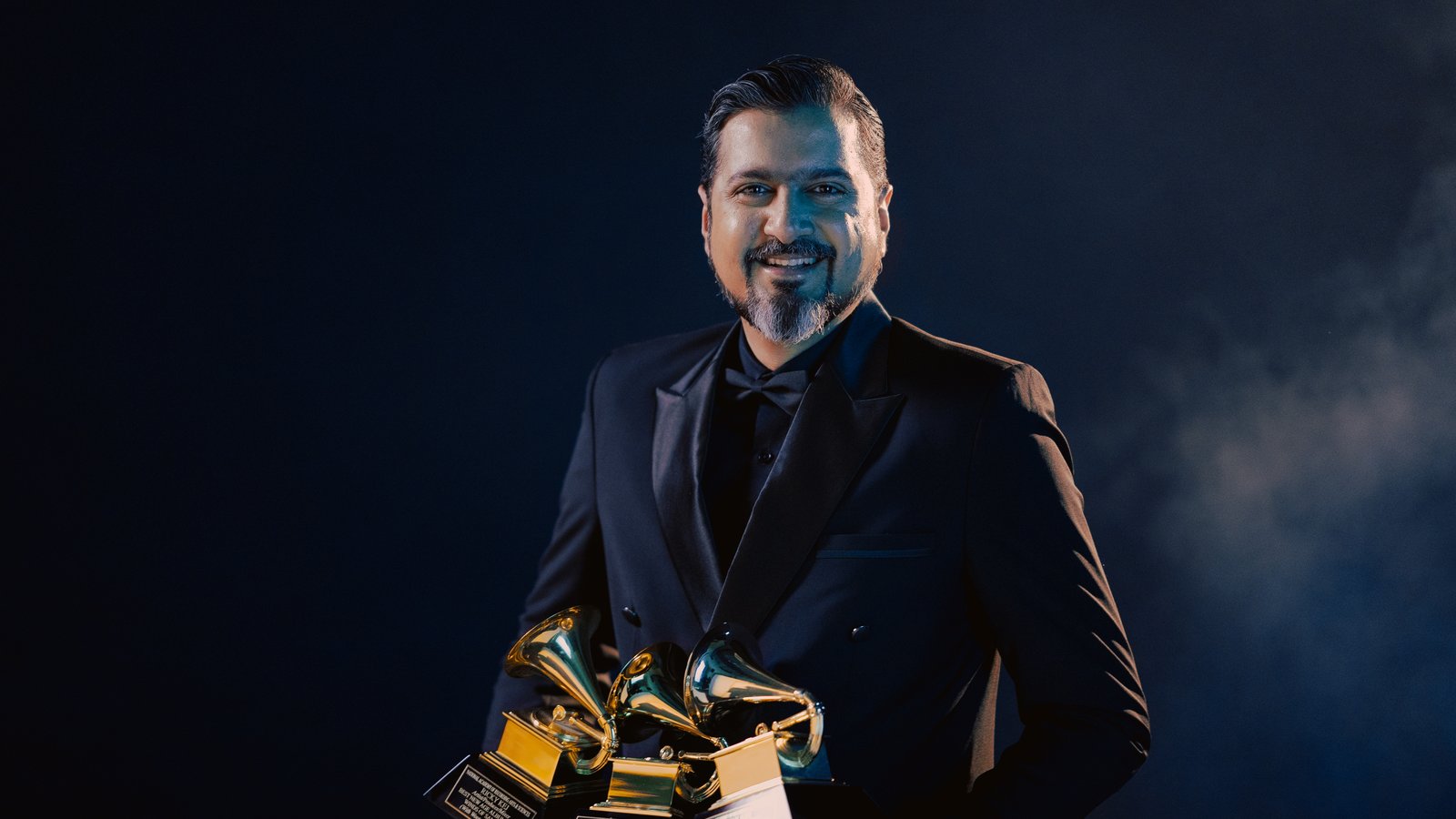 Composer Ricky Kej in a black tuxedo holding three Grammy Award trophies against a dark blue background