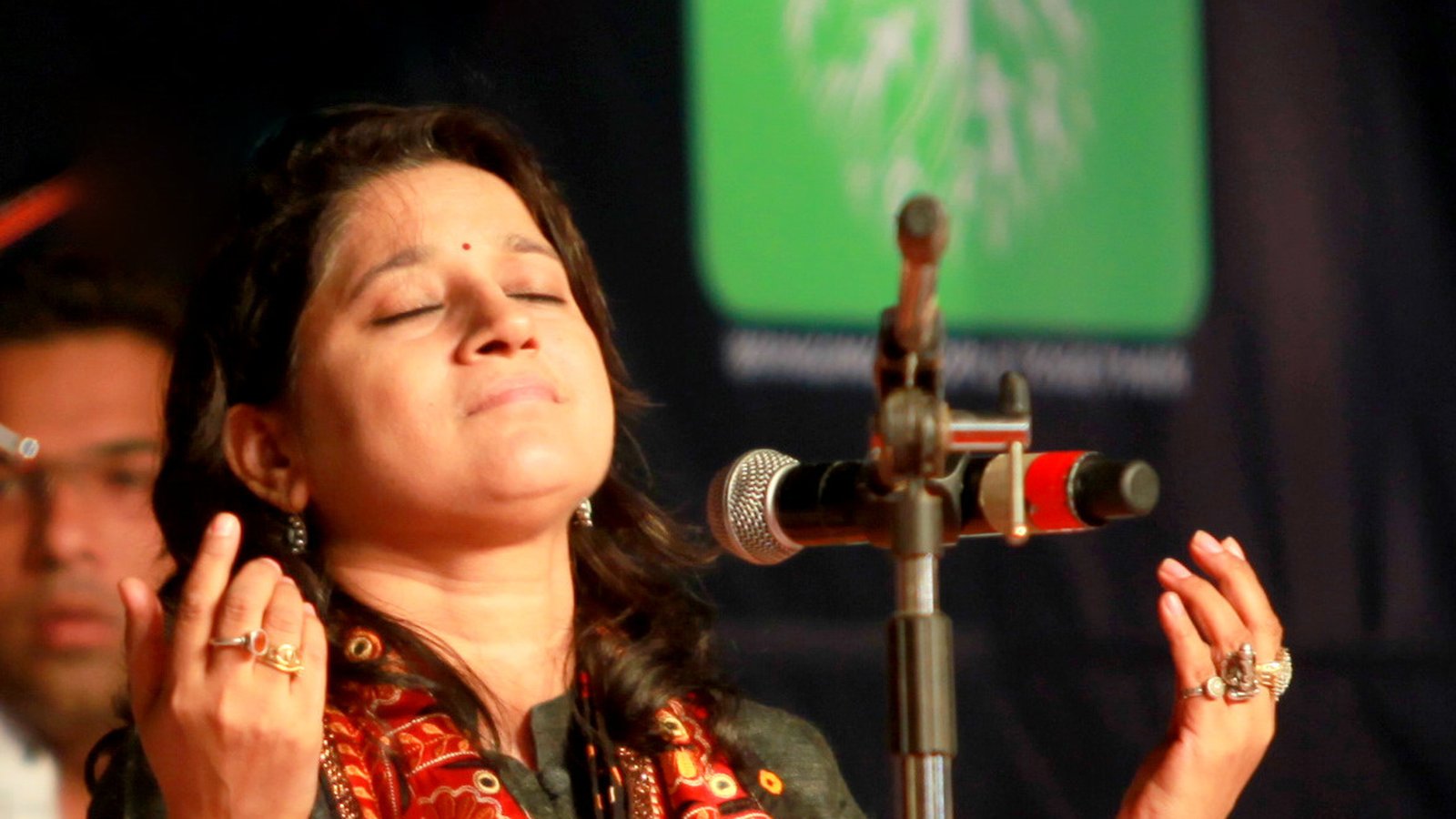 Kavita Seth singing with eyes closed and hands raised at a microphone during a Sufi concert, wearing a red embroidered stole